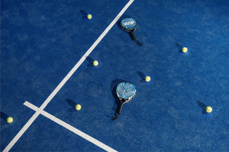 Bovenaanzicht van een blauwe padelbaan met witte lijnen, twee rackets en verspreid liggende gele ballen.