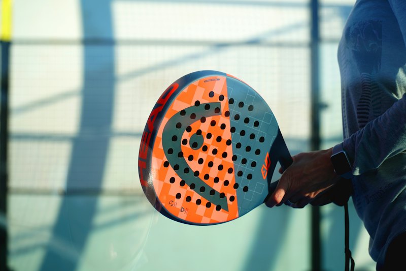 Een hand houdt een oranje-blauw padelracket vast, met de glazen wand van de baan erachter.